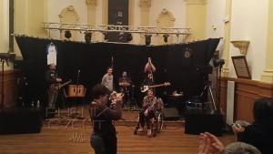 Témoignage de Patricia, présidente de l'association Sclérolife, avant l'ouverture du concert caritatif dans la salle de spectacle de la mairie de Fillières. Patricia est en fauteuil roulant, le groupe mickey trump est sur scène et écoute attentivement.