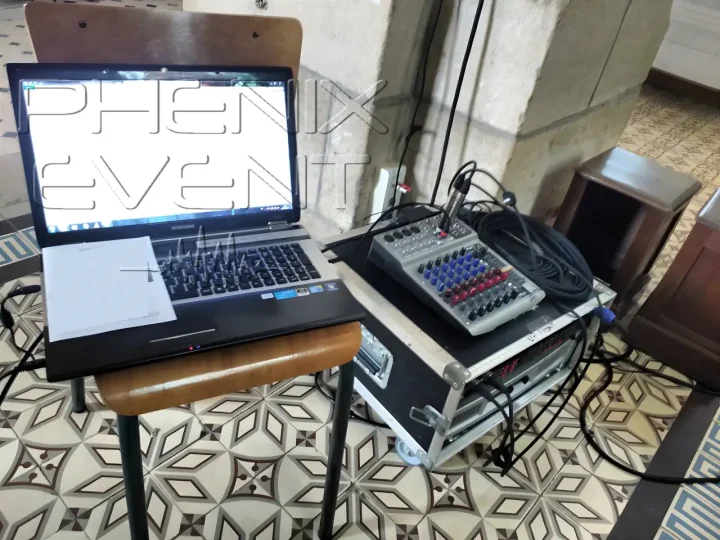 Rack d'amplification dissimulé derrière un pilier dans l'église, avec table de mixage et ordinateur posé sur une chaise